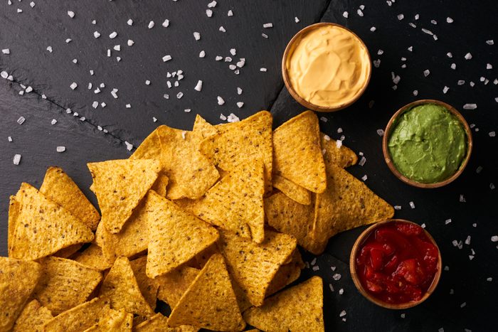Mexican nachos with sauces tomato ketchup, cheese and guacamole in wooden bowl on dark background, top view, copy space. Delicious salty corn chips triangular nachos snack for party