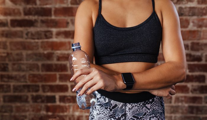 Mid section of a fit young woman holding water bottle against wall at fitness studio. Closeup of an athlete wearing a smartwatch relaxing.
