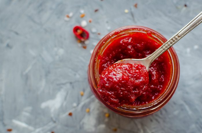 Red hot chili jam in glass jar with fresh ingredients on grey concrete background. 