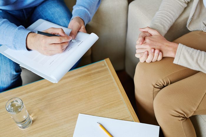 Female patient sitting on armchair and holding her hands together while male psychologist filling in medical card, close-up shot