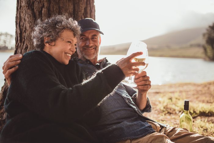Portrait of happy senior couple sitting near a lake toasting with wine on camping. Woman and man drinking wine at campsite.