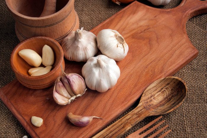 Heads of garlic, sliced garlic, and garlic cloves on a wooden cutting board, surrounded by wooden utensils like spoons and forks.