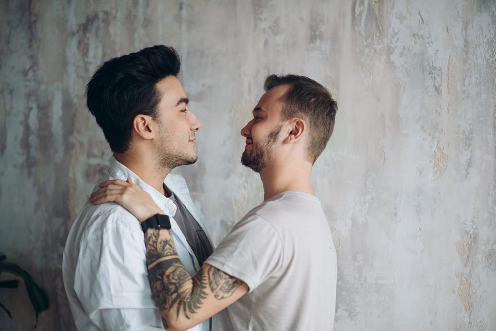 Hot gay couple at the sweet moment of love enjoying closeness and passion kissing isolated over grey lounge rustic background
