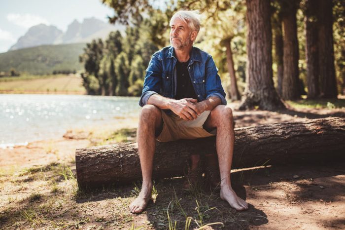 Portrait of a senior man sitting on a log near lake and looking away. Mature man sitting alone on lake.
