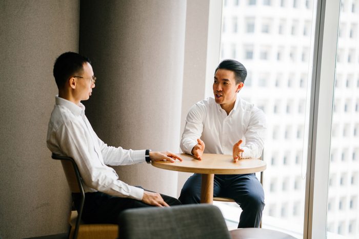 Two Chinese Asian business men have a chat at a table in the office in a meeting room during the day. They are professionally dressed in shirt and pants and having a focused discussion together.