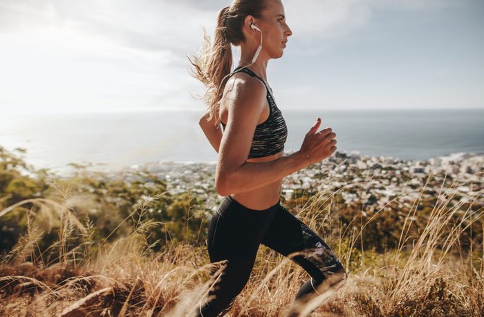 Side view of fit young woman wearing earphones running through mountain trail. Female runner training over rough trail in hillside.