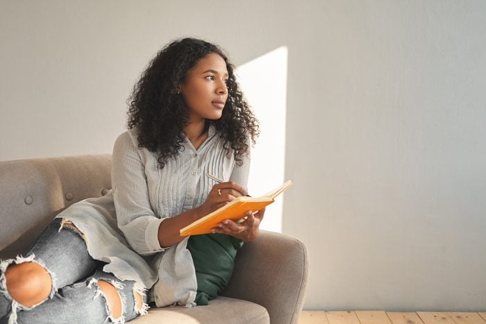 Attractive young dark skinned female with Afro hairstyle relaxing on couch at home, having pensive thoughtful look, writing down ideas for her own startup project, using pen and copybook