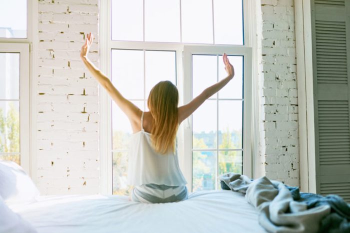 Woman stretching in bed after waking up, back view, entering a day happy and relaxed after good night sleep. Sweet dreams, good morning, new day, weekend, holidays concept