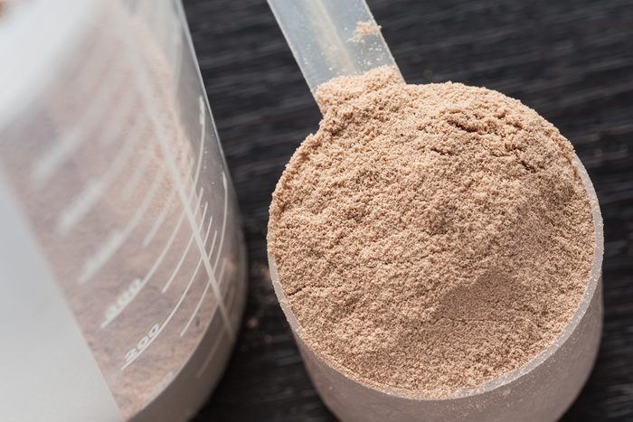 Scoop of chocolate whey isolate protein next to the translucent protein shaker, with focus on the protein inside the scoop