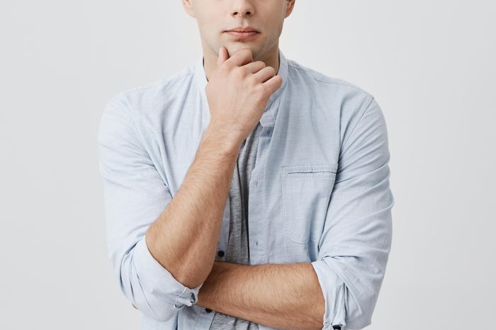 Puzzled thoughtful male student dressed in light blue shirt, holding hand under his chin, frowning face, looking upwards, being dissatisfied with problems at university, thinking over his mistakes.