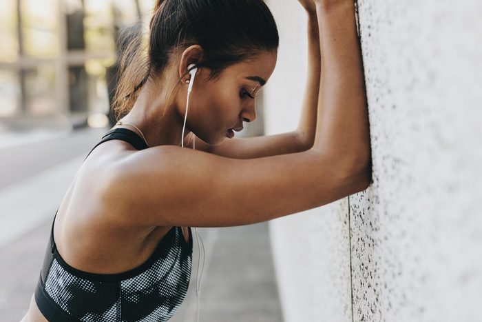 black woman in workout clothes, earbuds, leaning against a wall