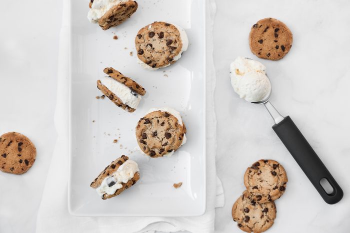 Chocolate chip cookie vanilla ice cream sandwiches with cookie crumbles all around. Ice cream scoop on the side with other cookies on a marble table top. Summer snacks.
