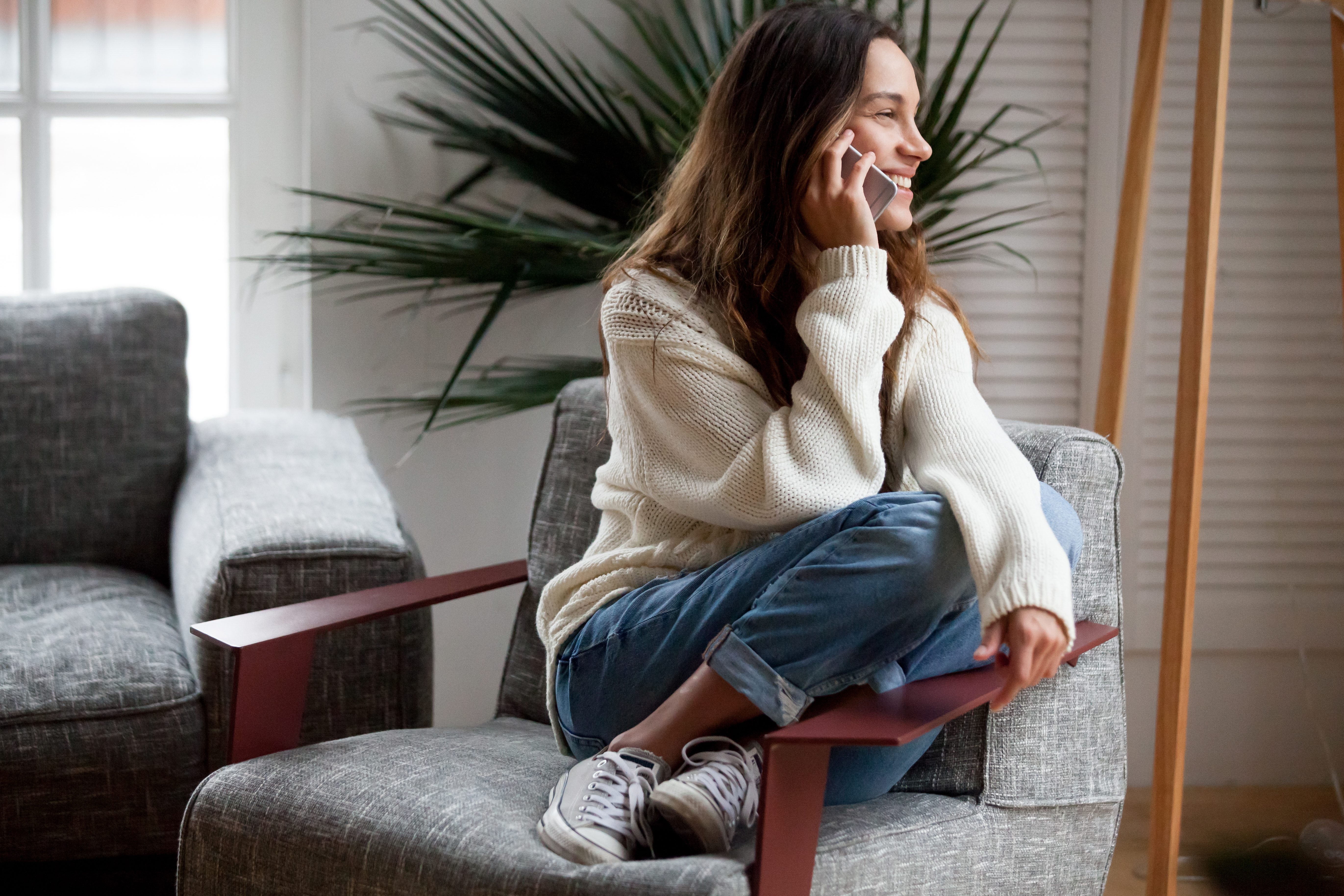 Happy cheerful young woman talking on the phone at home, smiling teen girl making answering call by cellphone sitting on chair, beautiful lady having pleasant funny conversation speaking by mobile