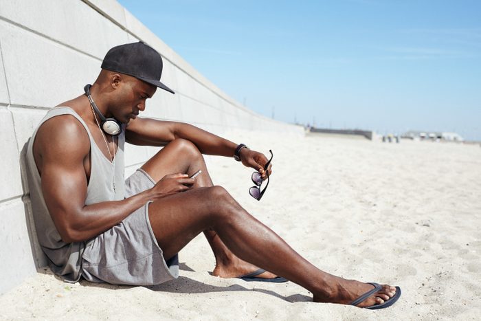 Young guy sending a text message at the beach. African man sitting on beach using mobile phone. Muscular male model outdoors.