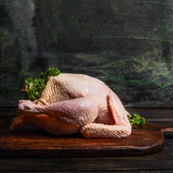raw whole chicken ready for cooking or roast on rustic kitchen table over dark background, side view.