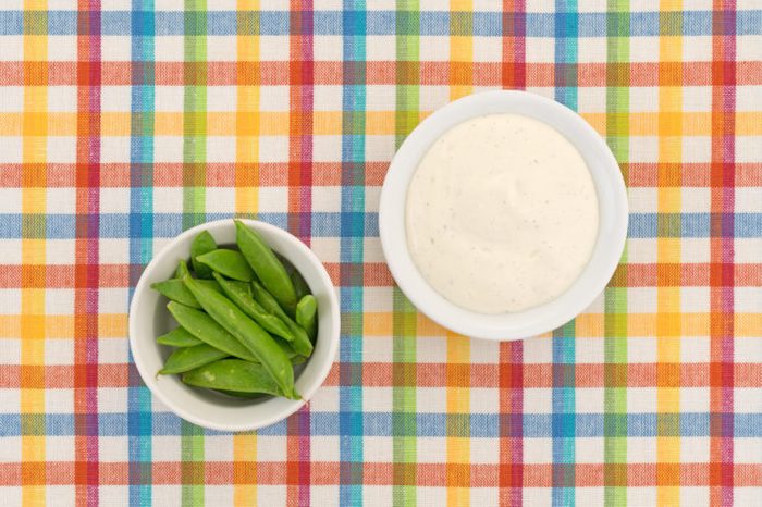 Top view of a small bowl filled with sugar snap peas and creamy ranch dressing to the side atop a colorful cloth patterned place mat.