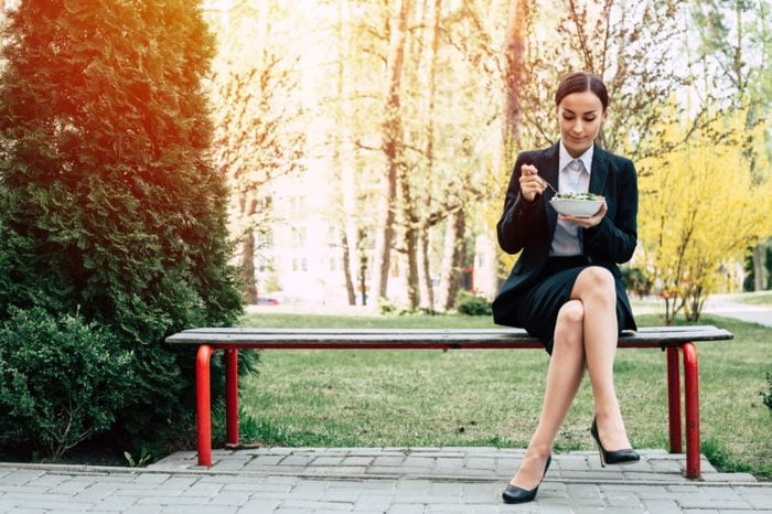 Modern beautiful business woman in a black business suit sits on a bench outside and eats a salad with fresh vegetables