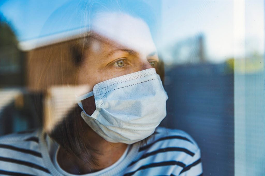 woman at home quarantine isolation looking out of window wearing a surgical mask