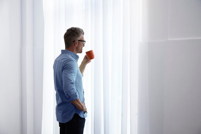 man standing in home looking out window and holding coffee mug