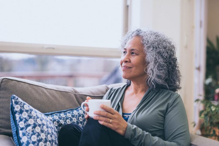 woman sitting on couch at home while holding a cup of coffee and looking out the window