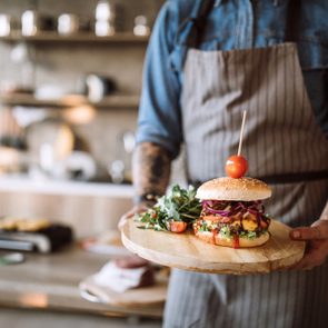 Man with apron standing in the kitchen and holding a wooden plate with prepared delicious hamburgers