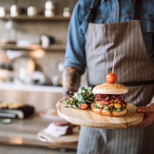 Man with apron standing in the kitchen and holding a wooden plate with prepared delicious hamburgers