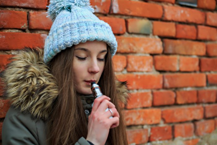 Vape teenager. Young pretty white girl in blue cap and green jacket smoking an electronic cigarette opposite brick wall on the street in the spring.