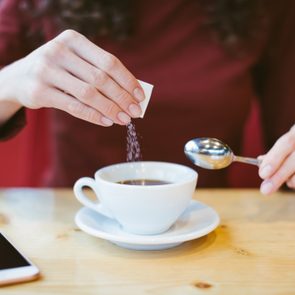 Woman's hands pouring sugar into black coffee - girl sitting at the table with espresso and smartphone - blood and glycemic index control for diabetes -excess of white sugar in food concept