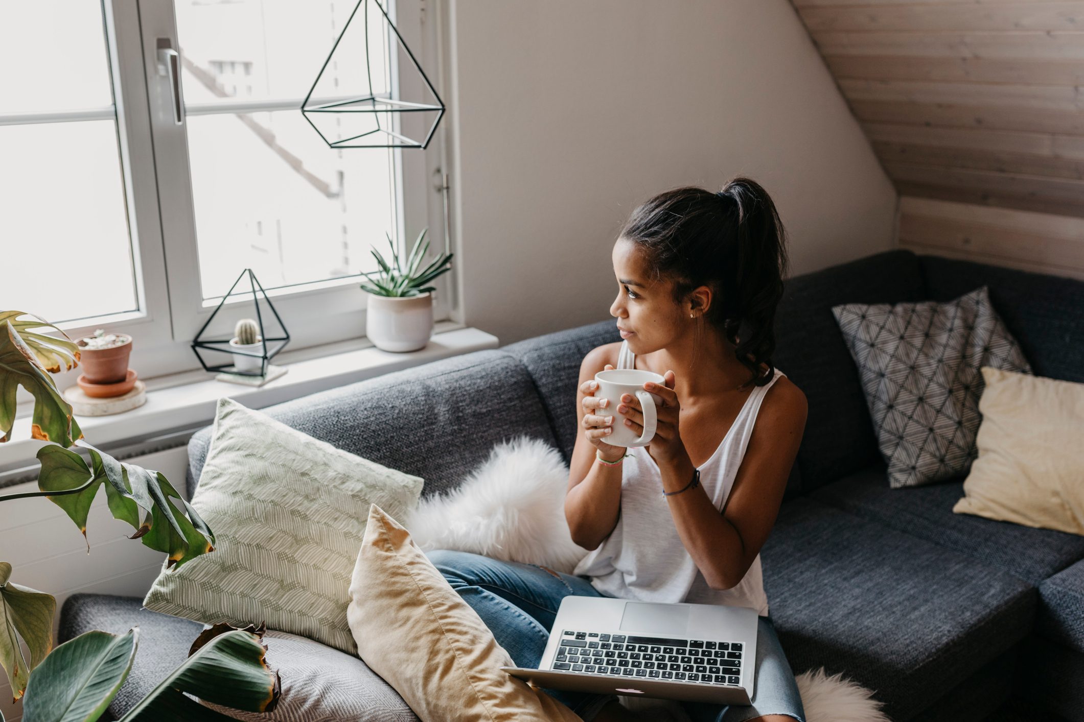 Young woman with laptop and cup of coffee sitting on the couch at home looking out of window