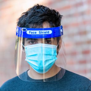 Young man posing wearing a face shield and a protective mask looking at the camera against a brick wall