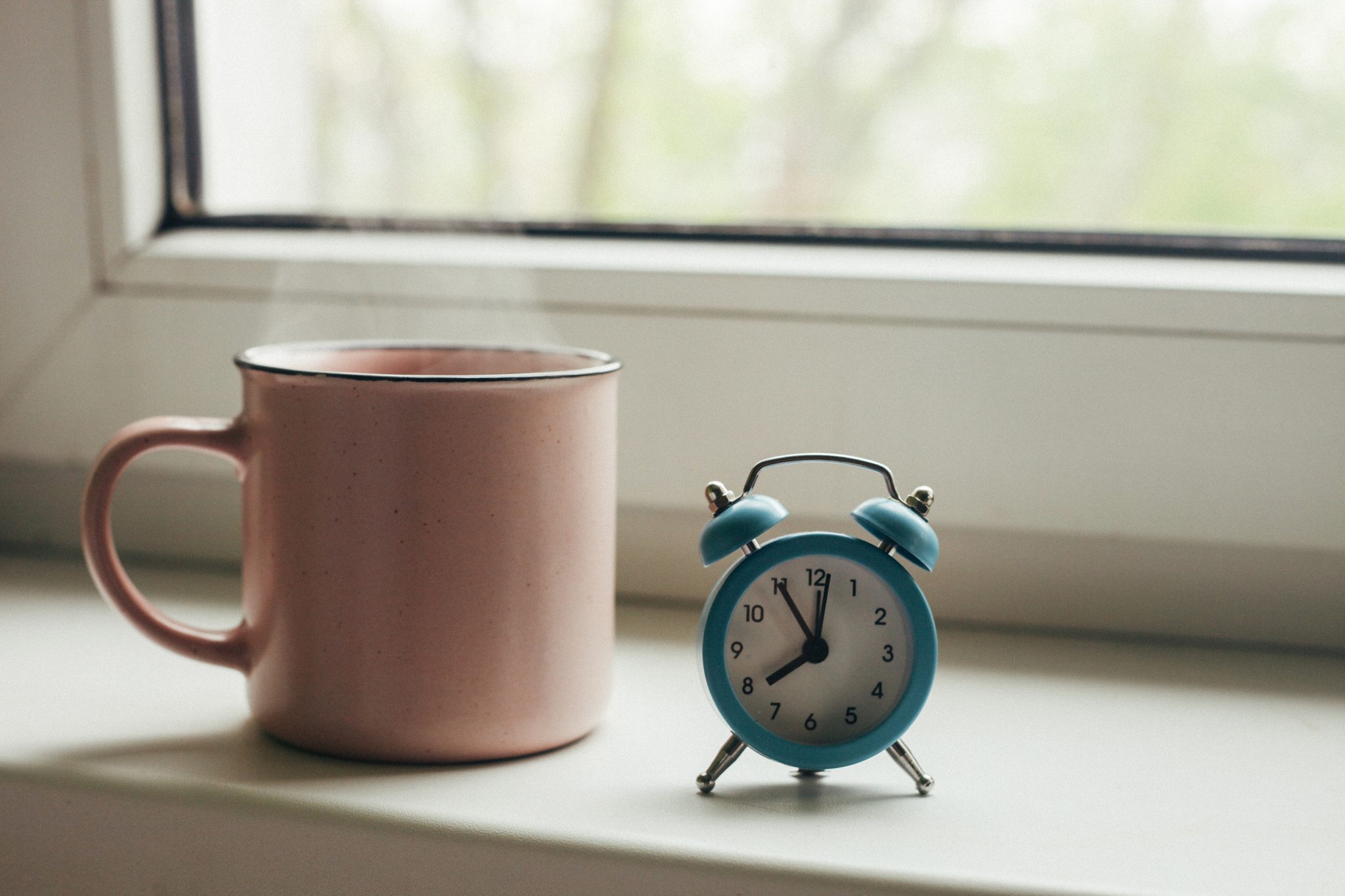 Pink cup of hot coffee with steam and retro alarm clock on window sill in morning
