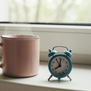 Pink cup of hot coffee with steam and retro alarm clock on window sill in morning