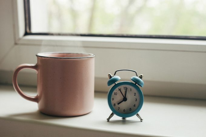 Pink cup of hot tea with steam and retro alarm clock on window sill