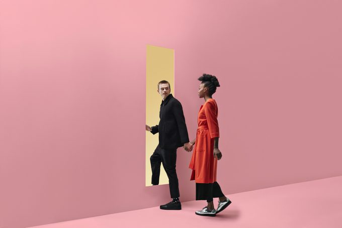 Woman & man holding hands, approaching rectangular opening in colored wall
