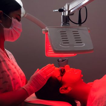 Young woman having red LED light facial therapy treatment in beauty salon. Beautician wearing face mask maintaining safety procedures during appointment. Beauty, new normal and wellness