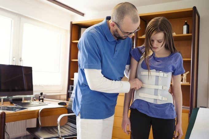 Male GP in a German medical office, assessing teenage girl wearing back brace for Scoliosis correction