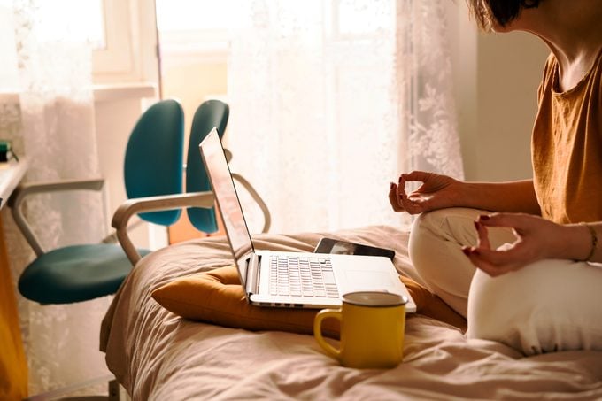 A middle-aged woman in white jeans and a yellow sweater sitting on the bed in a yoga pose in front of a laptop and a cup of coffee