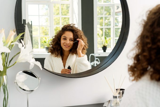 woman applying serum to face in bathroom mirror