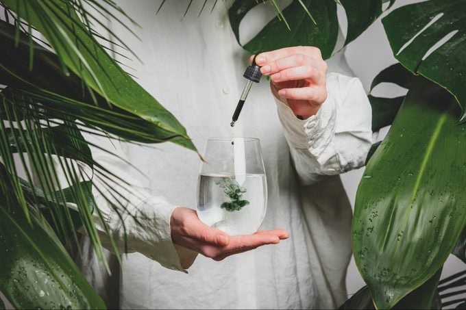 woman in a white shirt adds drops of chlorophyll to glass of water standing next to palm trees