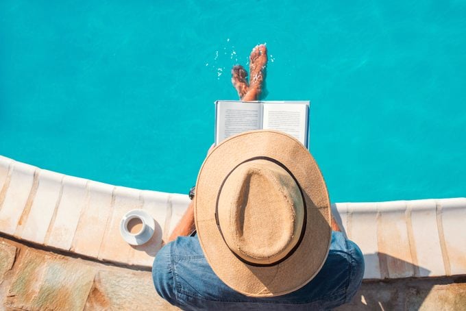 overhead shot of man wearing a hat and reading a book by the pool during summertime