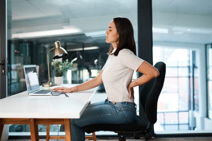 young woman suffering from back pain while sitting and working at desk in office