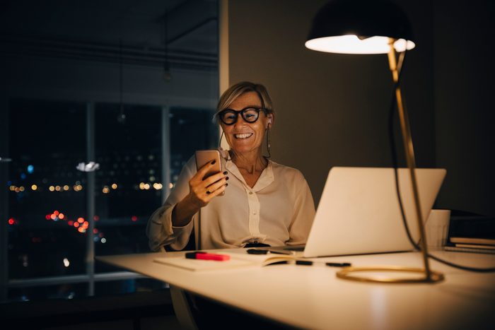 Smiling businesswoman using smart phone while sitting with laptop at illuminated desk in coworking space