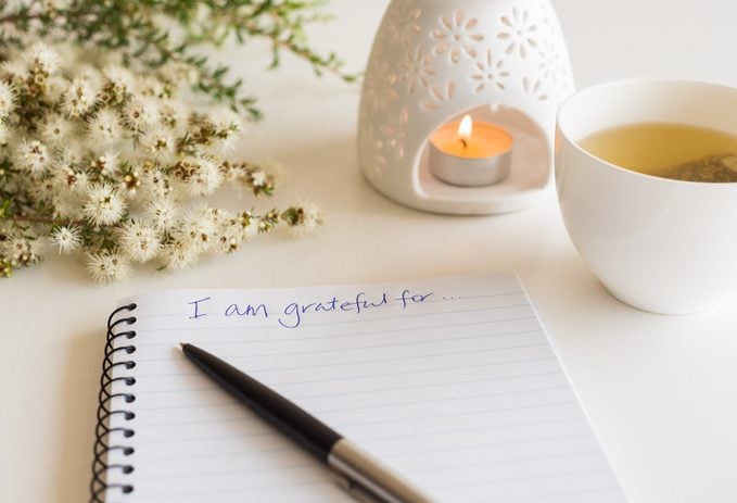 Close up of notebook with handwritten text "I am grateful for..." in foreground with pen, cup of tea, flowers and oil burner in soft focus