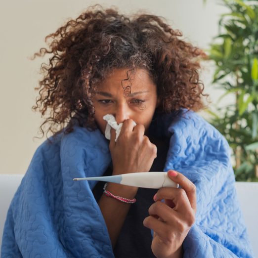 woman sick at home, wrapped in a blanket, holding a tissue to her nose with one hand and a thermometer in the other wondering if it's the flu, covid, or flurona