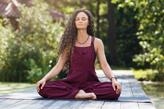 A beautiful woman in a burgundy overalls, practicing yoga, sitting on a mat in a lotus position, is engaged in meditation on the shore of a pond on a warm sunny morning