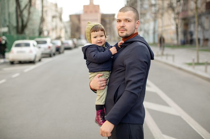 Young refugee man in the blue jacket stands on the road. He holds his baby daughter carefully and looks into the camera.