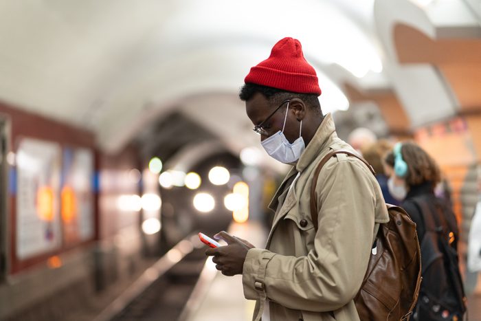 Black Man Waiting For Train At Subway Station, Using Mobile Phone, Wear Face Mask. Covid 19 Pandemic