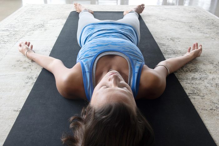 woman laying down doing yoga inside on a yoga mat, the corpse pose