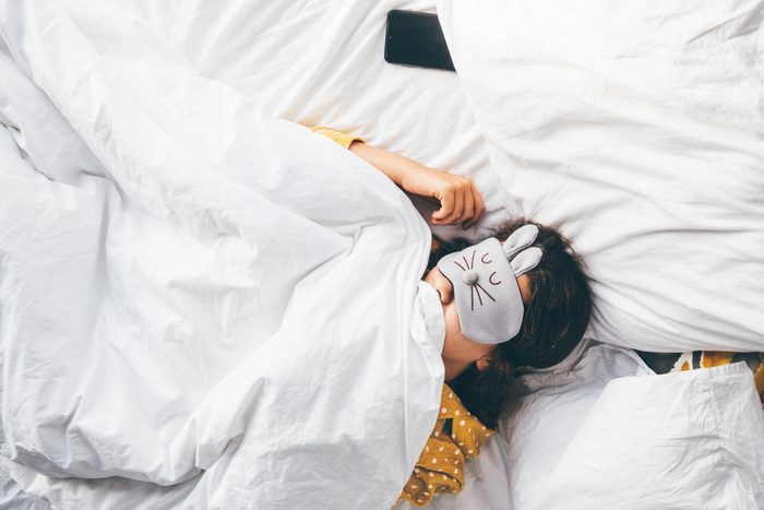 Happy millennial woman lying in bed with sleeping mask and phone in a white bed