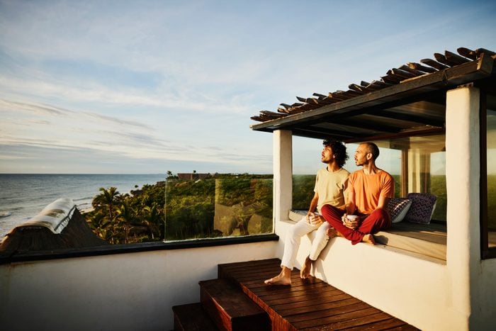 Wide shot of gay couple sitting on rooftop deck of luxury tropical beachfront villa while drinking coffee and watching sunrise